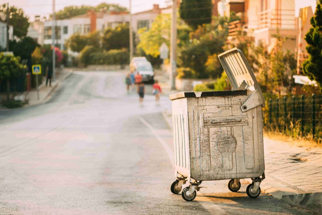 Metal Waste Container On Wheels In Street During Summer Sunny Evening.